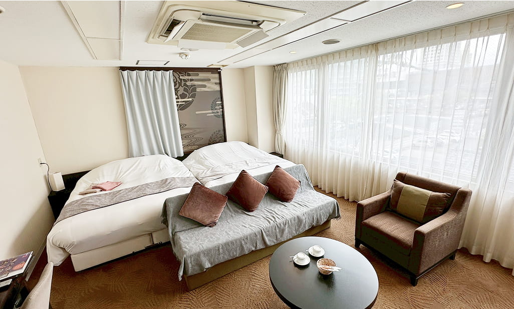 大きな窓から自然光が差し込む、ツインベッドとソファが配置された明るいホテルの客室