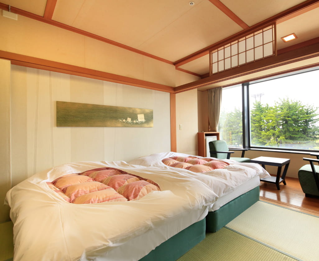 畳の上にベッドが置かれた和モダンな客室で、大きな窓から緑の景色を望める