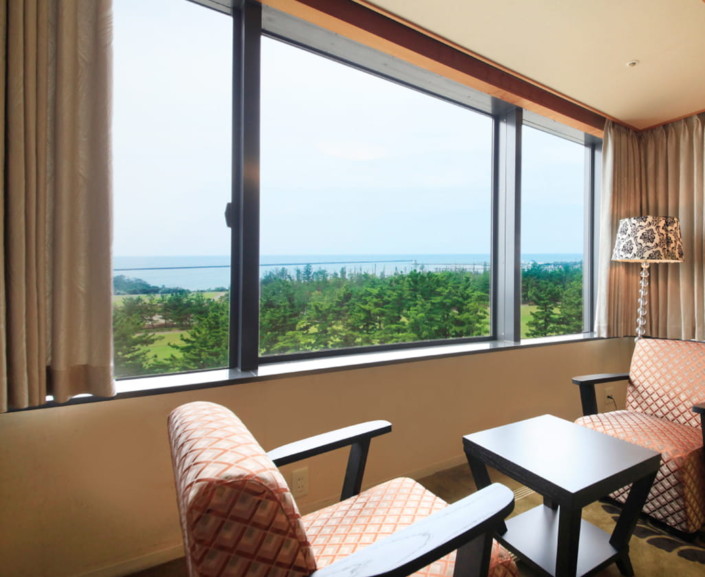 窓際にチェアとテーブルが配置された洋風のくつろぎスペースで、海と森が見渡せる絶景の眺望